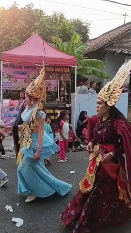 The video shows a parade or procession on a street in what appears to be a village. In the foreground, a woman in an elaborate red and gold costume with a large headdress walks past a food stall. Behind her, a woman in a blue hijab and a striped shirt walks. Spectators, including children and adults, are seated along the side of the street watching the procession. In the background, houses and trees are visible, and some stalls are set up. The overall atmosphere seems festive.