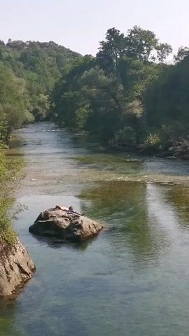 A person is lying on a rock in the middle of a river. The river is clear and shallow, with a rocky bottom visible. Trees and foliage line the banks of the river, and a hill covered in trees is in the background. The sky is bright and clear, indicating a sunny day. The person is wearing a blue shirt and red shorts. They appear to be relaxed and enjoying the scenery.