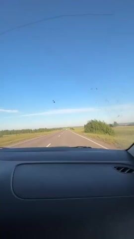 The video is filmed from inside a car, looking through the windshield. The windshield has several cracks and is splattered with dirt or insects. The car is driving on a paved road that stretches into the distance. The road is bordered by flat, grassy fields and scattered trees on both sides. The sky is clear blue with a few wispy white clouds. The overall scene depicts a car traveling through a rural landscape on a sunny day.