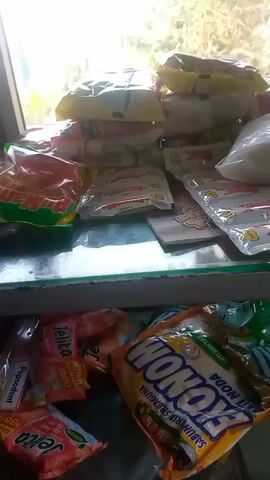 The video shows a close-up view of several products displayed on shelves. On the top shelf, there are various packaged goods, including what appear to be food items and cleaning supplies. Some packages are brightly colored with text and images. On the bottom shelf, more products are visible, including packets of "Jelita" and a large orange and yellow package labeled "WONOKO SABUN CUCI". The lighting suggests an indoor environment, possibly a store or market stall, with natural light coming from the background. The overall impression is a neatly arranged display of retail items.