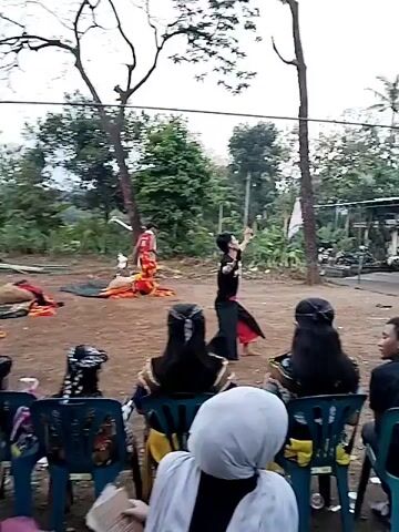 The video shows a man performing a traditional dance outdoors. The man is wearing a black shirt and black pants with a red sash.  He is barefoot. He is performing in front of a group of people sitting in chairs.  The people in the chairs are wearing traditional clothing. Behind the dancer and to the left is another person in a red and yellow costume.  The background includes trees, a dirt ground, and a building.
