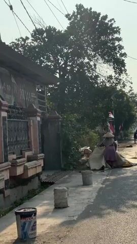 A person wearing a conical hat and a purple skirt, and a grey headscarf is partially visible, pulling a large beige tarp. Next to them, there are logs and a white tarp. A bag of rice is on the ground, along with other bags. The scene appears to be outdoors, with trees and houses in the background. Indonesian flags are visible, suggesting a celebration or national event. The ground is paved with a mix of asphalt and dirt. Several buckets are also present.