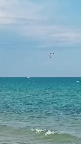 The video shows a clear blue sky with a few white clouds above a turquoise ocean. In the distance, a kiteboarder is visible on the water, with a colorful kite in the air. The waves are gentle, and the overall scene is peaceful and serene. There are a few other distant boats visible on the horizon. The kiteboarder is performing some kind of maneuver or riding on the water.