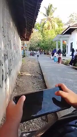 A first-person perspective video captured by someone holding a smartphone, likely while riding a motorcycle given the slight motion blur and the handlebar visible at the bottom of the frame. The person is filming a street scene. On the left is a rough, dirt path adjacent to a concrete wall with graffiti. On the right is a paved walkway leading to a white building with a light blue trim. Several people, predominantly women and girls wearing headscarves, are walking along the paved walkway, some in groups. In the background, there are trees, including a palm tree, and more people are visible further down the street, as well as parked motorcycles. The lighting suggests it is daytime, possibly late afternoon due to the angle of the sun and the shadows. The person holding the phone appears to be interacting with the device, with their thumbs positioned on the screen.