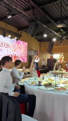 The video shows a celebration or event taking place indoors, possibly at a banquet hall or reception venue. In the foreground, a round table is set for a meal with white tablecloths, plates of food, glasses, and drinks. Several people are seated around the table, appearing engaged and joyful. One woman in the center is smiling and raising her hand.

In the background, there is a stage area with a large screen displaying a vibrant, colorful image featuring silhouettes of people with raised arms and floral or abstract designs. A man in a vest and bow tie is on the stage, possibly performing or speaking, as he gestures with his hands. Another person, dressed in a white shirt, stands to the left of the stage. To the right of the stage, a cluster of balloons is visible.

The overall atmosphere seems festive and lively. The lighting suggests it is likely evening or indoors with artificial lighting. The ceiling structure with exposed beams and lighting equipment indicates a functional event space. While a specific date or time is not explicitly shown, the large screen might be displaying an event title or theme.

The location context provided is Vyshhorod, Ukraine, but this is not visually discernible from the video itself. The visible text on the screen is primarily graphic and not easily decipherable as legible words, though the number "2026" is faintly visible as part of the large graphic. The people's expressions and body language convey a sense of celebration and enjoyment.