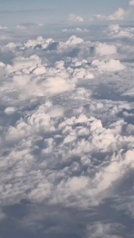 The video shows an aerial view of clouds. The clouds are white and fluffy, with some areas of darker gray. The camera is moving slowly, giving the impression of flying through the clouds.