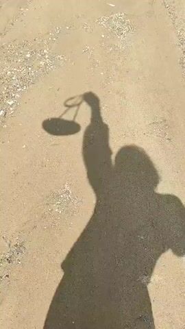 The video shows a shadow of a person holding a phone or camera up, likely taking a selfie. The person's shadow is cast on a sandy or dirt ground with small pebbles. The shadow shows the person wearing a coat and boots, with a bag or purse strap over their shoulder. The scene appears to be outdoors in daylight. The person's shadow moves slightly throughout the video as if they are walking or adjusting their position.