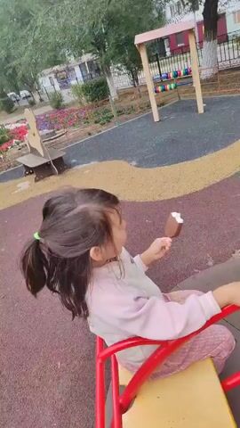 A young girl with dark hair and a white and pink outfit is sitting on a yellow and red merry-go-round, eating an ice cream bar. She is looking towards the camera and smiling. The merry-go-round is on a playground with a red, yellow, and dark grey rubberized surface. In the background, there is a swing set, a seesaw, and a building. The weather appears to be overcast.