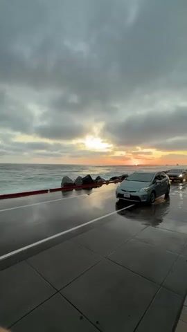 The video shows a coastal road during a cloudy sunset with waves crashing onto the road. Several cars are driving on the wet road. A person on a bicycle rides past a light blue car that is partially submerged in water. Another car is visible in the background.