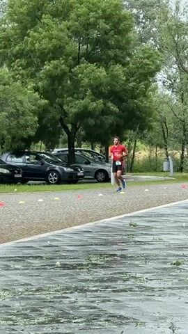 Here's a description of the video:

The video shows a man running in a park-like setting, possibly during a race or training session. He is wearing a red shirt and black shorts, and blue running shoes. He passes by a photographer who is kneeling down to take pictures. A second person, wearing a brown hooded sweatshirt and blue pants, stands nearby looking at a phone or similar device. Cars are parked along the edge of the area, and trees provide a green backdrop. There are orange and white cones marking a path, and the ground appears to be wet, possibly from rain.
