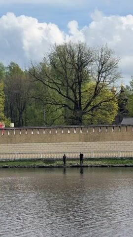The video shows an Orthodox church situated behind a stone wall on the bank of a body of water, possibly a river or a lake. The church is white with dark roofs and golden domes topped with crosses. The wall is tan with regular decorative stone elements along the top. Two figures stand along the bank in front of the wall, possibly fishing. The sky is partly cloudy, and trees are visible in the background, showing early spring foliage. The water is calm, reflecting the scene above. The camera pans slightly to the left, revealing more of the church.