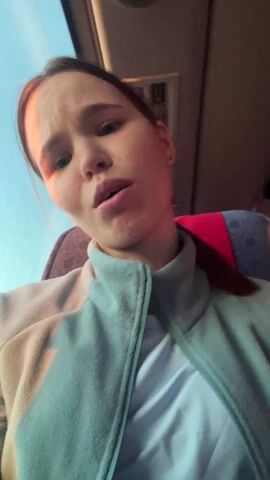 A young woman is recording herself, presumably via her phone. She's seated in a bus seat with a red headrest, wearing a light blue fleece jacket and a white t-shirt. The bus window is visible to her left, showing a clear, bright sky outside. She is singing along to a song playing in the background and appears to be expressing emotion through her facial expressions. The bus interior is visible but blurry in the background.