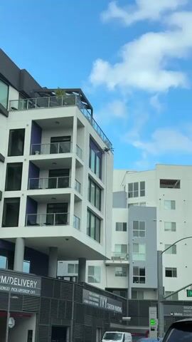 The video shows a high-rise building on a partly cloudy day. A drone is cleaning the windows of the building, spraying water down the glass. Below the building is a parking garage and a sign that reads "Ivy Station Parking". There is also a sign that reads "Ramen/Delivery" with "pick-up/drop off" underneath. A white van is parked in front of the garage.