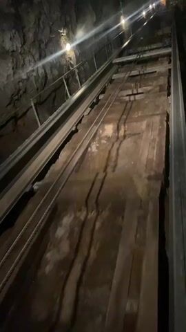 The video shows a POV shot of a funicular ascending an inclined tunnel. The tunnel walls are rough and rocky, illuminated by artificial lights spaced along the ceiling. The funicular track consists of two metal rails on the left and a wooden track on the right. Thick cables run parallel to the track. The funicular itself appears to be a wooden platform with metal support beams and wheels. The platform is slowly moving upwards, indicated by the changing perspective and the blur of motion. No people are visible in the video.