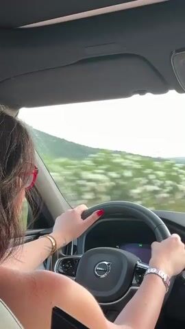 The video shows a close-up view from inside a car, looking forward. A person's hands are on the steering wheel of a Volvo. The person has red nail polish and is wearing bracelets. The car is moving, as indicated by the blurred scenery outside the windows. The scenery consists of green hills and some pink flowers along the side of the road. The sky is a light blue with some clouds. A dashboard is visible in front of the steering wheel.