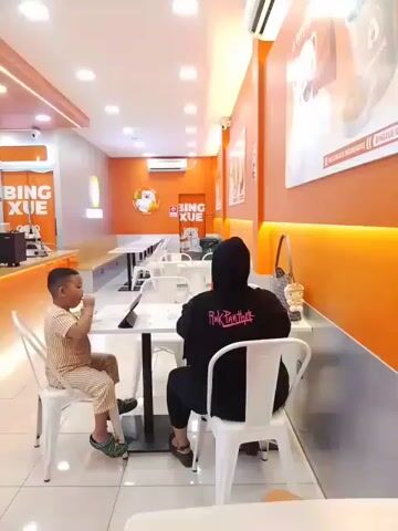 A young boy in a striped outfit sits at a white table in a brightly lit establishment with orange walls. He is eating something with his hands and looking at a tablet on the table. To his right, a person wearing a black hooded garment is seated at the same table, facing away from the camera. Behind them, a counter area is visible with various machines and displays, including a large screen showing a promotional advertisement. The establishment appears to be a cafe or dessert shop, indicated by the branding "BING XUE" on the walls and a display of what looks like ice cream or drinks. The flooring is tiled, and the ceiling has recessed lighting. The overall atmosphere is clean and modern.
