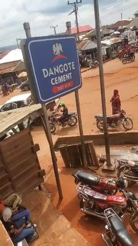 The scene depicts a street corner in a bustling town. A sign indicating the direction to Dangote Cement stands prominently. Numerous motorcycles are parked nearby. People and motorcycles traverse a dusty, reddish-brown road. The area is surrounded by various buildings and structures, including stalls and houses. Overhead, power lines intersect the sky. Two people sit casually at the base of a building.