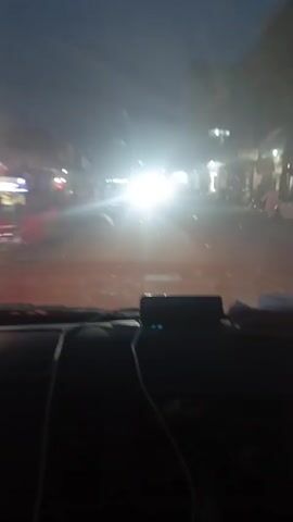 The video shows a view from inside a vehicle, likely a car, driving down a street at night. Streetlights and headlights of oncoming vehicles create a bright glare. Buildings line the street, and some people are visible walking on the sidewalks. The dashboard of the car is visible in the foreground. The overall atmosphere is dimly lit with bright points of light. A siren can be heard.