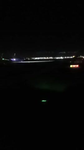 From an interior, seated perspective at night, blurred lights of an airport tarmac and structure are visible in the distance through a window.  A wing of the plane in which the person is seated passes in front of the window at the beginning of the video.
