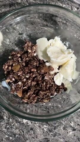A close-up shot shows milk being poured from a jug into a clear glass bowl. The bowl contains granola, some raisins, and scoops of vanilla ice cream. The milk mixes with the granola and ice cream, creating a creamy texture. The background is a dark gray, speckled countertop.