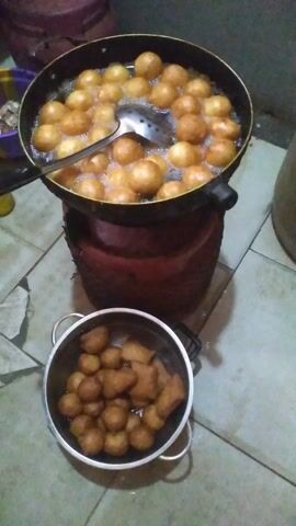 The video shows a top-down view of a cooking scene. In the foreground left, a black frying pan filled with golden-brown fried dough balls is immersed in hot oil. A silver slotted spoon is resting in the pan, scooping some of the dough balls. To the right of the frying pan, a metal colander holds a batch of fried dough balls, some of which are round and others are more rectangular in shape. The dough balls appear to have just been fried and some are still glistening with oil. In the background, a large red plastic container is partially visible. The floor beneath the cooking setup is tiled. The lighting suggests an indoor setting, possibly a kitchen, with artificial light sources. The focus is on the food being prepared.