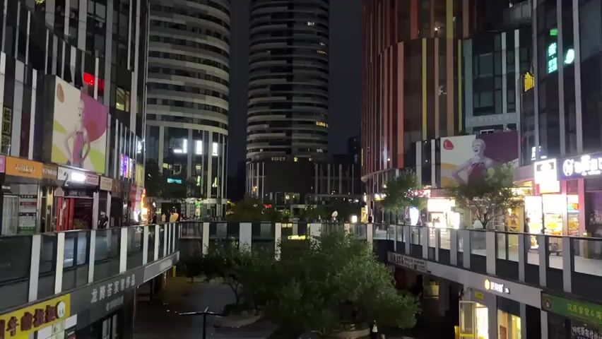 The video shows a nighttime view of a modern urban plaza. Tall buildings with distinctive facades, including one with vertical colored stripes and two cylindrical structures, dominate the background. Elevated walkways with railings connect the buildings, and shops with illuminated signs are visible on multiple levels. A few trees are planted in the central area, and scattered individuals are seen walking on the walkways and in the plaza below. The lighting suggests artificial illumination from the buildings and signs, creating a vibrant yet somewhat subdued atmosphere. The scene appears to be a commercial district or shopping area in a city.