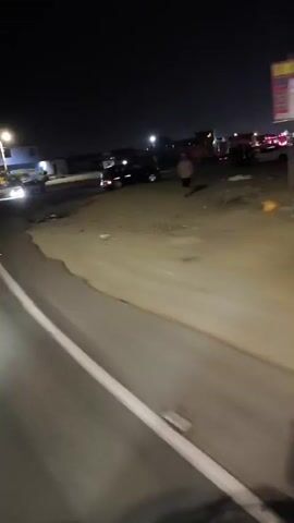 At night, the camera moves past a multi-vehicle accident scene. A black SUV is positioned in the center of the area, while a white police truck with flashing red lights is parked to the left. A severely damaged white car is visible further to the right, with several individuals standing near it. Debris is scattered across the ground. The scene is illuminated by headlights and streetlights. The road surface is asphalt, with a white line marking the edge. Silhouetted figures, presumably including the camera holder, appear in the foreground. The sky is dark.