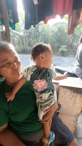 An elderly woman in a green shirt holds a baby dressed in a white and green outfit with a pink flower embellishment. The baby is facing to the right, with their left hand extended outwards. The woman is looking slightly downwards and to her left. In the background, there are clothes hanging, foliage, and a road with a vehicle passing by. The scene appears to be outdoors, with sunlight illuminating the area.
