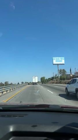 The view from inside a moving vehicle looking ahead at a highway under a clear blue sky is shown. A white tractor-trailer truck is moving in the same direction in the center lane. A blue billboard advertising insurance is to the right of the road. There is a grey guardrail on the left side of the road. A second vehicle is briefly visible on the right side of the frame. The dashboard of the recording vehicle is visible at the bottom of the frame.