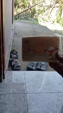 A hand holds a small package with cartoon rabbits on it. The package is held in front of a doorway. To the left of the doorway, a pair of sandals are placed on the ground. To the right of the doorway, two pairs of flip-flops are visible. Beyond the doorway, a dirt path leads to trees and foliage. The ground in front of the doorway is tiled.