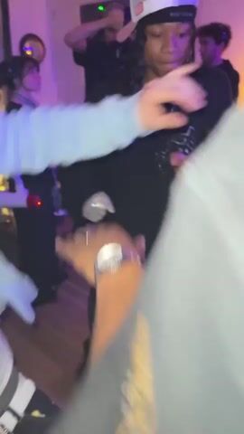 The video captures a lively indoor scene where a person with dreadlocks and a white baseball cap, wearing a black sweater and matching pants, is dancing energetically. They are surrounded by other people, one holding a white cup, and another with a watch visible. A cameraman is filming the scene, with lighting adding to the ambiance. The flooring appears to be wooden, and the overall setting looks like a casual gathering or party.