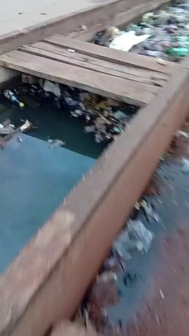 The video shows a long, open concrete drainage ditch alongside a road. The ditch is full of a dense layer of trash and debris, including plastic bags, bottles, and other unidentifiable waste. In some sections, there is stagnant water beneath the trash. Wooden planks are placed across the ditch at intervals, likely serving as makeshift bridges. The road runs parallel to the ditch and has vehicles and pedestrians visible in the background. The overall impression is one of neglect and poor sanitation.