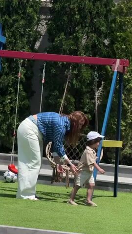 A woman with long brown hair, dressed in a blue and white striped long-sleeve shirt and light-colored wide-leg pants, stands on artificial green grass next to a swing set. She is facing towards a macrame hanging chair swing, with her back partially to the camera. A young child, wearing a beige short-sleeved shirt, beige shorts, and a white baseball cap, approaches the swing set from the right, extending their right arm slightly. The swing set has red and blue metal poles, holding two swings: the macrame chair and a red plastic swing. A white and black soccer ball rests on the grass to the left of the frame. Tall green foliage and a light-colored wall are visible in the background under clear daylight.