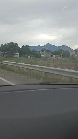 This video shows a view from inside a vehicle, looking out at a road, a metal guardrail, and a rural landscape. The sky is overcast and gray. In the background, there are trees, some low-lying buildings, and mountains in the distance. A traffic sign indicating a T-junction is visible. One of the buildings has signage that appears to be in a foreign language. The overall impression is a somewhat bleak, overcast day in a rural or semi-rural area.