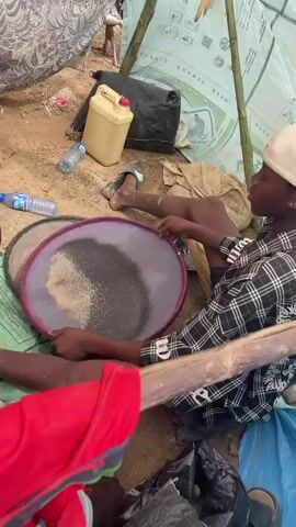 A person sits in what appears to be a makeshift tent, holding a circular pan filled with a mix of dark and light material, possibly sand or sediment. They are wearing a plaid shirt and a white cap. There is a second pan nearby, along with bottles of water, a yellow container, and other miscellaneous items scattered on the ground. The tent is constructed from bamboo poles and covered with a tarp.