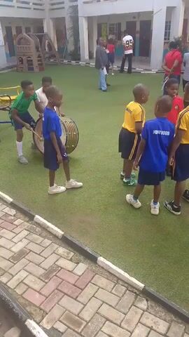 A group of young children, dressed in school uniforms, are gathered on a green artificial turf area in what appears to be a schoolyard. Some of the children are holding musical instruments, including a large bass drum and sticks. One boy in a blue shirt and dark shorts is walking towards the viewer. To the left, two boys in green and white shirts are standing near the bass drum, with one boy holding drumsticks. Several other children in blue and yellow shirts are grouped together further back, some appearing to be facing the drum. In the background, there is a multi-story building with visible windows and balconies, and other adults and children can be seen near the entrance of the building. The area is bordered by a brick walkway with a black and white striped edge.