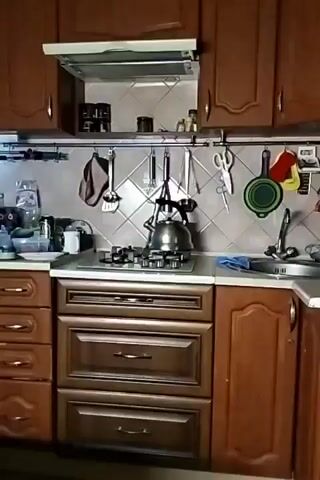 The video shows a kitchen with wooden cabinets and drawers. Above the countertop is a range hood, and below is a gas cooktop with a kettle on it. Various cooking utensils are hung on a rack. On the left side of the countertop are kitchen appliances and items such as a blender, thermos, and containers. A sink is visible on the right side. The camera slowly pans from right to left, showing the entirety of the kitchen setup.