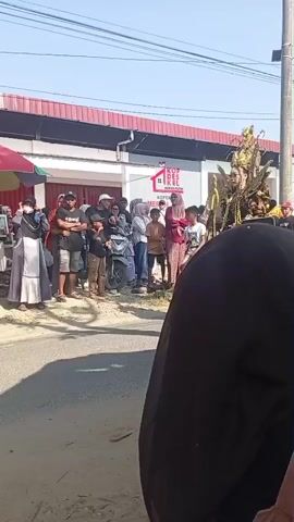 The video shows a street scene with a crowd of people watching a parade or procession. In the center of the frame, a person in an elaborate costume, possibly a performer or a float, is walking down the street. The costume appears to be golden and black, with intricate details and a long train. Several people in the crowd are holding up their phones to record the event. The background shows buildings and a clear blue sky. There are also some parked motorcycles. The scene suggests a festive or cultural event taking place outdoors.