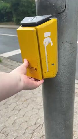 A person's left hand is shown pressing a yellow pedestrian crossing button mounted on a metal pole. The button has a white icon of a hand holding a cane, indicating it is for pedestrians with mobility impairments. The button is located on a sidewalk next to a street. In the background, there is a traffic light showing a red pedestrian signal, trees, buildings, and a car.