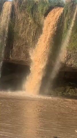 The video showcases a waterfall in Kukum, Nigeria. The water, a muddy brown color, initially cascades down a rocky, stepped cliff face into a wider body of water below. The cliff is predominantly light brown, with horizontal striations and patches of green vegetation on its surface and along its top edge. As the video progresses, the flow of water significantly decreases, eventually stopping almost entirely, leaving only a few trickles down the cliff. The water body at the base reflects the brown hue of the falling water. The scene appears to be under clear, daylight conditions.