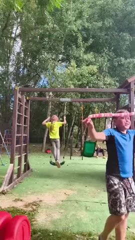 A boy in a blue t-shirt and patterned shorts stands on a playground holding a pink and white striped toy snake above his head. His mouth is open, and he appears to be laughing. A girl in a yellow t-shirt and gray leggings is on a swing in the background. The playground has swings, a climbing structure, and green turf. Trees and foliage are visible in the background, and the sky is blue with some clouds. The boy is in the foreground, slightly to the right of the center of the frame. The girl is to the left of the frame.