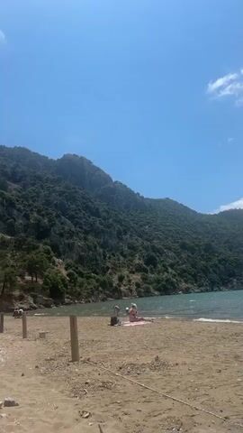 The video shows a beach with people and a large, tree-covered mountain in the background. The sky is blue with some clouds. The beach has light-colored sand and some driftwood or wooden posts. There are a few people sunbathing or playing near the water. The mountain is covered in dense green vegetation. The video pans slightly to the left. A rock barrier is visible in the foreground.