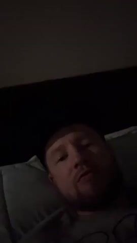 A person is lying down with their head on a pillow. The person is looking at the camera and making a peace sign with their right hand. The person has short hair and a beard. The person is wearing a gray shirt. The background is dark and out of focus.