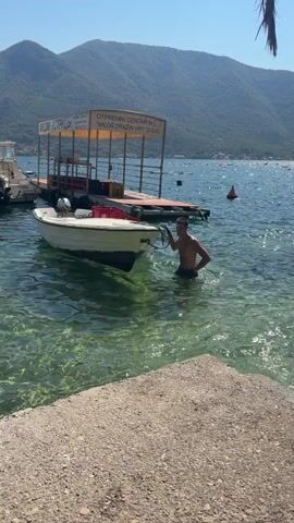A young man is standing in clear, shallow water near a boat. He is holding a rope attached to the front of the boat and appears to be moving it. The boat is white with a small outboard motor on the back. Behind the boat, there is a wooden dock with a covered structure on it. The dock has signs with text in a foreign language. In the background, there are mountains and other boats on the water. The sky is clear and blue. The sun is shining. The man is wearing dark-colored swim trunks.