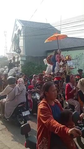 A group of people are participating in a parade or procession on a street. The procession includes individuals in traditional clothing, some carrying decorative floats on their shoulders, which appear to be shaped like mythical creatures or animals. Some of the floats are adorned with umbrellas. Several people are riding on motorcycles, some following behind the procession and others to the side, observing or participating in the event. The environment appears to be a residential area with buildings and some greenery visible. The sky is bright, suggesting daytime.