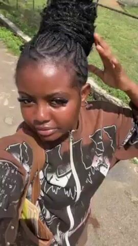 A young woman is standing on a paved surface outdoors. She is fair-skinned, with dark eyes, dark hair styled in braids and an updo, and large false eyelashes. She is wearing a brown and black long-sleeved shirt and carries a brown handbag on her shoulder. The woman smiles and gives a peace sign gesture to the camera. The background includes grass, a metal fence, and some buildings in the distance. The lighting appears to be bright daylight.