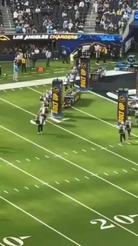 The video shows a marching band walking on a green football field. They are dressed in uniforms of white, blue and black. They are marching in line on a green field marked with white lines and numbers.  On the sides of the band are black pillars with the words "BOLT UP" in yellow. In the background is a crowd seated in a stadium under the sign "Los Angeles Chargers."