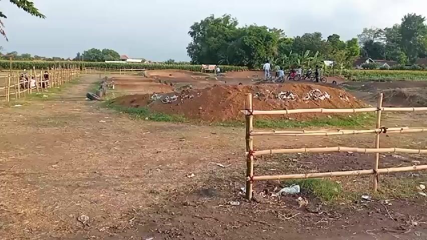 The video shows an outdoor dirt track with several mounds of dirt, some possibly for jumps. A bamboo fence with red ties separates the foreground from the track. In the background, there are trees, some crops, and a few houses. On the track, a group of people, some with bicycles, are gathered near the dirt mounds. The sky is overcast.