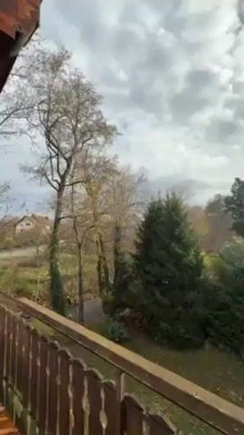 The video shows an outdoor view from a balcony. The sky is cloudy with a few glimpses of sunlight. There are trees and houses in the background. The balcony has a brown railing. There are some bins in view at the bottom of the frame.