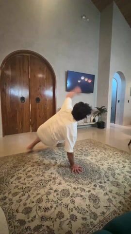 A man with dark curly hair wearing a white t-shirt and khaki shorts is breakdancing on a patterned rug in a living room. The man is executing a series of acrobatic movements, supported by his hands and occasionally his head. The living room has textured light gray walls, a high ceiling, and a polished concrete floor beneath the rug. In the background are two arched doorways, a large wooden double door, a flat-screen television displaying various circular icons, and several potted plants. A blue cushion is visible in the foreground.